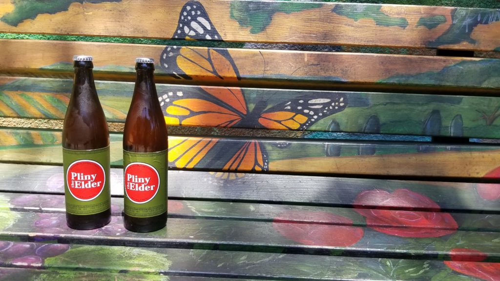 Two bottles of Pliny the Elder on a park bench. 